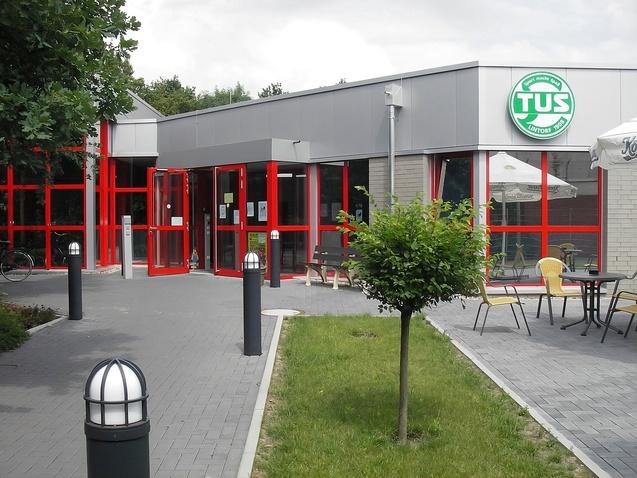 Eingangsbereich eines sportlichen Vereins mit roten Fenstern und einem grünen Logo, umgeben von einer gepflegten Außenanlage.