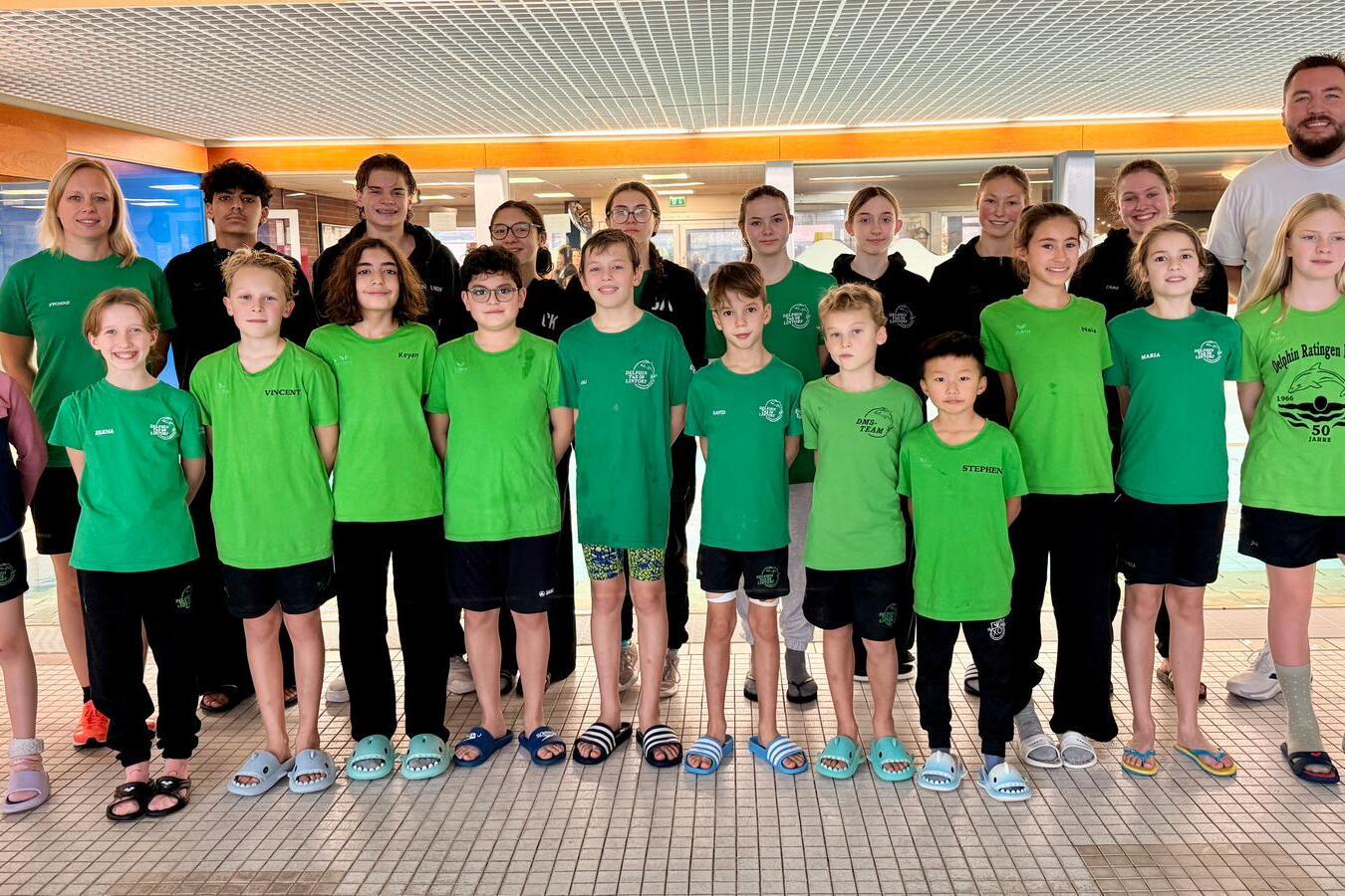 Gruppenshot von Schwimmern in grünen Shirts vor einem Schwimmbecken, begleitet von Trainern und Betreuern.