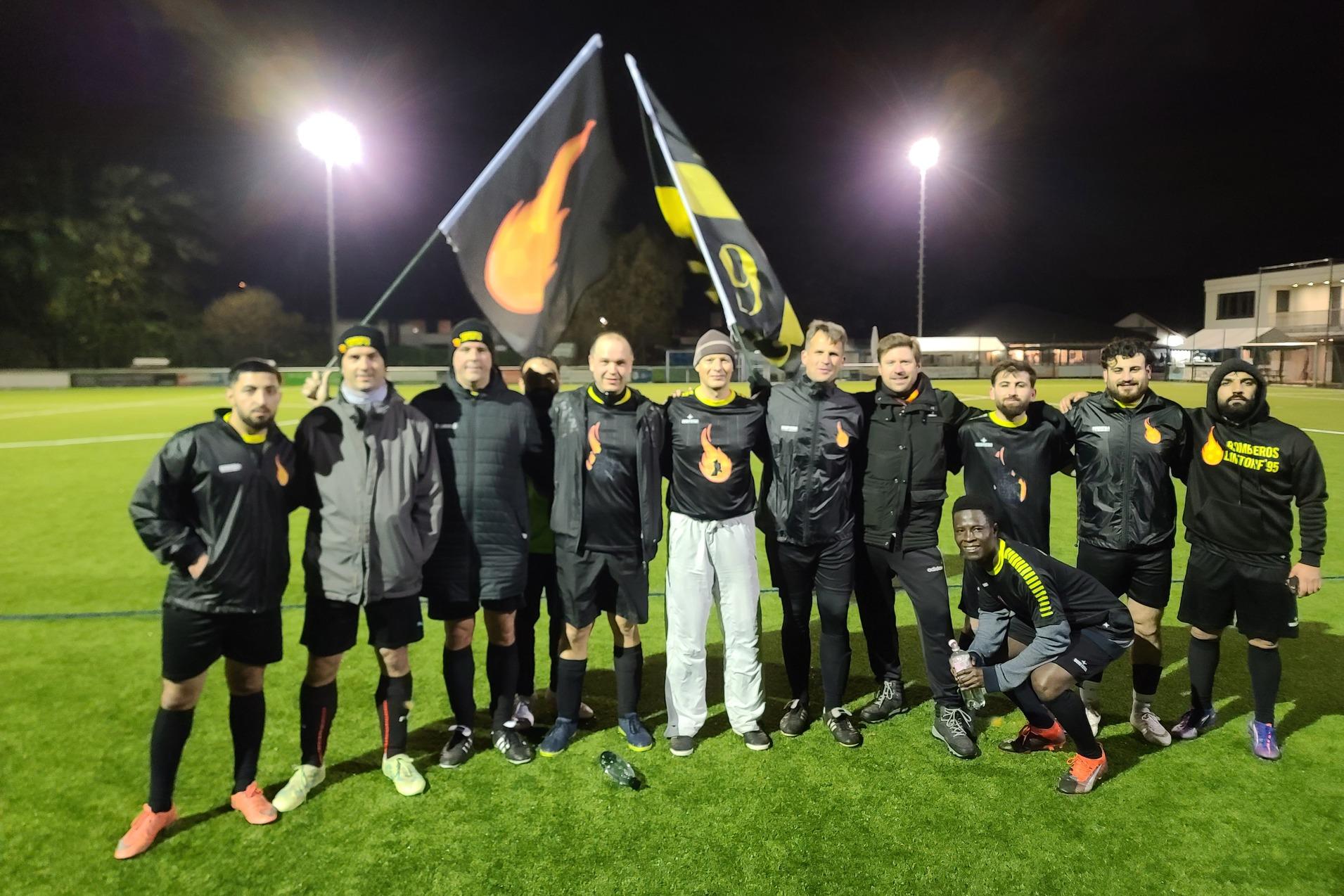 Gruppenshot von 11 Fußballspielern in einheitlicher Sportkleidung, die Flaggen auf einem beleuchteten Feld schwenken.