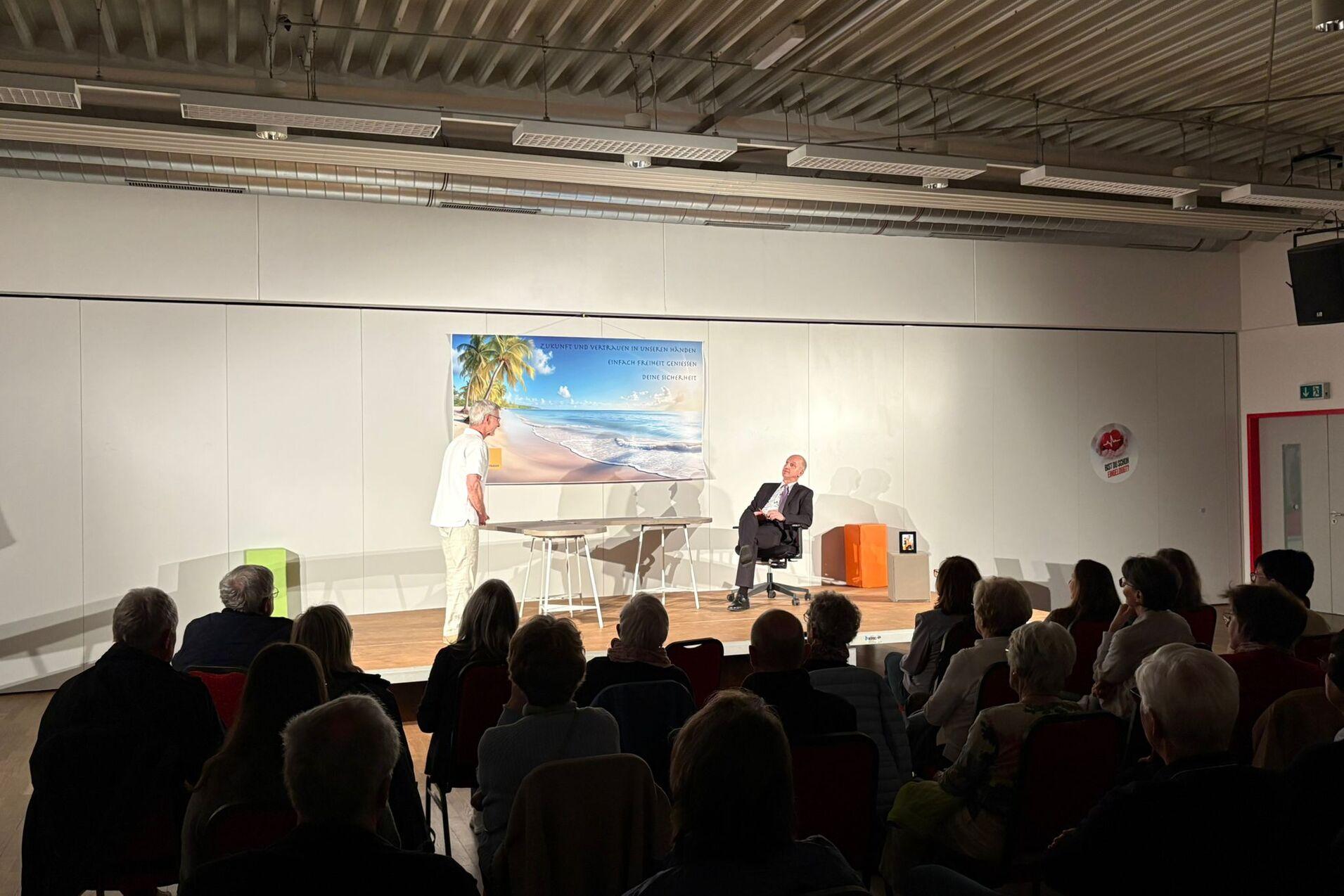 Zwei Personen auf einer Bühne, einer spricht, während Publikum in Stuhlreihen zuhört. Hintergrund mit Strandmotiv.